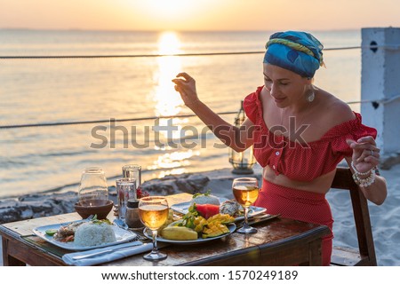 Young woman having romantic dinner in hotel restaurant during sunset near sea waves on the tropical beach, close up. Girl is enjoying the sunset with a glass of wine. Concept of leisure and travel