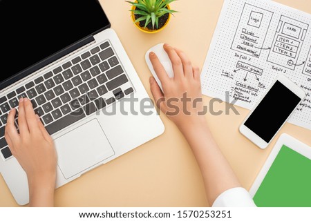 partial view of designer using laptop near gadgets, website design template and green plant on beige background