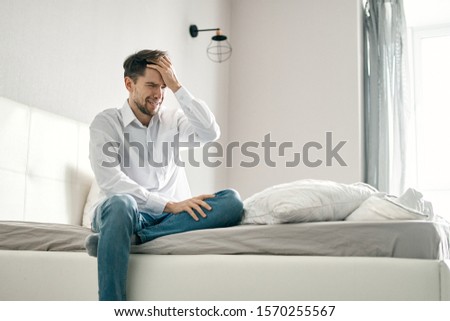 Jeans white shirt. A handsome man sits on the bed and touches his head with his hand