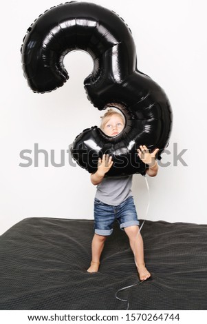 Portrait of toddler holding big black number balloon in his hands. Standing on bed. Inside. White background, not isolated.