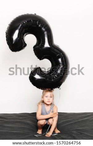 Portrait of sad toddler sitting on bed and holding big black number balloon in hand. Blonde hair, blue eyes. Indoors. White background, not isolated.