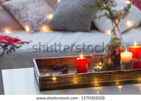 Composition of candles with juniper in vase on  white table against the background of  sofa with plaids and pillows. Cozy home concept