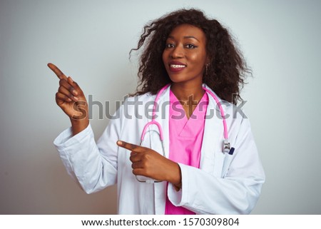 African american doctor woman wearing  pink stethoscope over isolated white background smiling and looking at the camera pointing with two hands and fingers to the side.