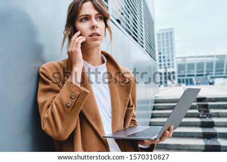 Young serious businesswoman in coat talking on cellphone working on laptop outdoor