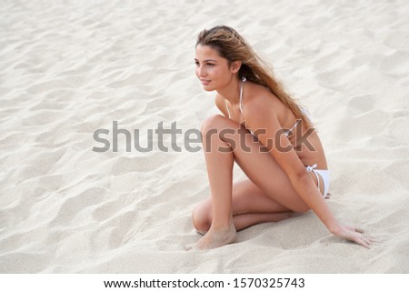Beautiful young woman in white bikini sitting on beach sand dunes, serene contemplative, relaxing on summer holiday nature space outdoors. Healthy female beauty body on vacation, recreation lifestyle.