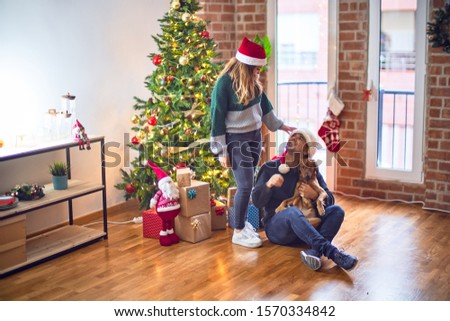 Young beautiful couple smiling happy and confident.. Sitting on the floor wearing santa claus hat hugging dog around christmas tree at home