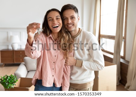 Happy young family couple holding key to new home on moving day concept, first time real estate owners man husband embrace woman wife look at camera proud buying property stand in own flat with boxes Royalty-Free Stock Photo #1570353349