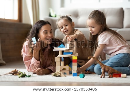 Happy young mum babysitter helping playing game with little kids daughters on floor, female nanny mother and cute small children sisters building castle of wooden blocks having fun with toys at home Royalty-Free Stock Photo #1570353406