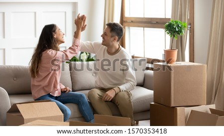 Happy proud millennial couple give high five celebrate family goal achievement moving into new home, young family roommates tenants renters owners sit on sofa with boxes relocate in own house concept Royalty-Free Stock Photo #1570353418