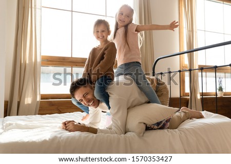 Funny cute kids daughters sit on fathers back on bed, happy little children girls having fun with cheerful dad playing horse piggyback game laugh in cozy sunny bedroom enjoy family morning activity