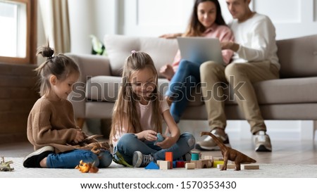 Happy little kids siblings preschool daughters having fun playing toys on floor while parents sit on sofa using laptop in comfy living room, family relaxing at home enjoying cozy leisure lifestyle Royalty-Free Stock Photo #1570353430