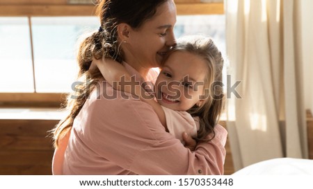 Caring young mum hugging cute sweet little daughter enjoying tenderness at home lit with sunlight, happy family mother and small child kid girl embracing cuddling at home, love in motherhood concept
