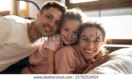 Happy family young father mother and funny little child daughter relaxing bonding lying on bed looking at camera, cute small girl with parents having fun cuddling enjoy morning in bedroom, portrait