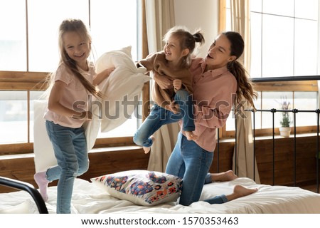 Happy family mom and two little cute children daughters having fun pillow fight on bed, carefree adult mother laughing play funny game with small kids siblings in bedroom enjoy morning life together