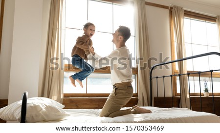 Funny cute kid daughter laughing jumping on bed playing with dad in bedroom interior, happy father holding lifting small child girl flying in single daddy arms enjoying family morning life having fun