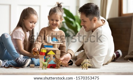 Caring young dad babysitter and cute small kids daughters playing with dinosaurs and wooden blocks sit on floor, father enjoying teaching helping little children holding toys having fun at home