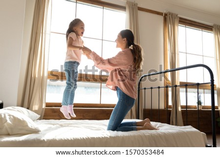 Cute active little kid girl jumping on bed flying in air playing with mum, happy parent young mother having fun with funny small child daughter laughing enjoying leisure morning together in bedroom