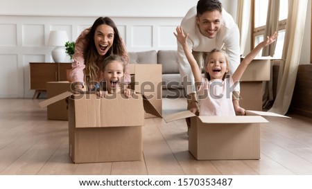 Excited young family having fun on moving day, happy parents pushing cardboard boxes with funny cute kids daughters riding in containers looking at camera laughing celebrate relocation into new home Royalty-Free Stock Photo #1570353487