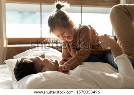 Adorable cute little kid daughter laughing tickling playing with dad on bed on sunny morning, happy father relaxing having fun with funny small child girl bonding enjoying leisure together in bedroom
