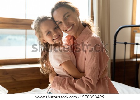 Portrait of happy affectionate family young adult mum embracing cute pretty small kid girl on bed, loving mother with little funny daughter hugging cuddling looking at camera in sunny morning bedroom