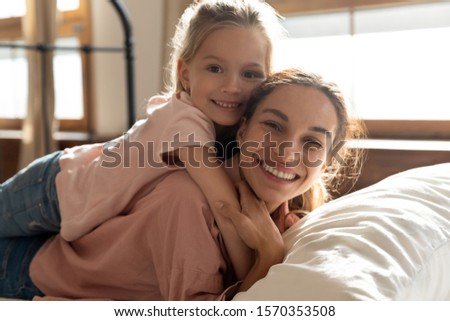 Adorable little girl embracing mom lying on her back on bed looking at camera, happy beautiful family young foster care parent single mother and cute small daughter hugging in bedroom, portrait