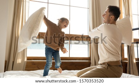 Excited funny small kid daughter playing pillow fight with dad on bed, happy cute little child girl and young father having fun in bedroom enjoying family leisure game morning activity together