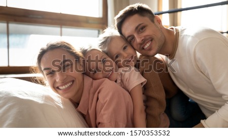 Affectionate beautiful family young adult parents mom dad and cute small children kids daughters bonding lying on bed in bedroom lit with sunlight looking at camera enjoying family morning, portrait