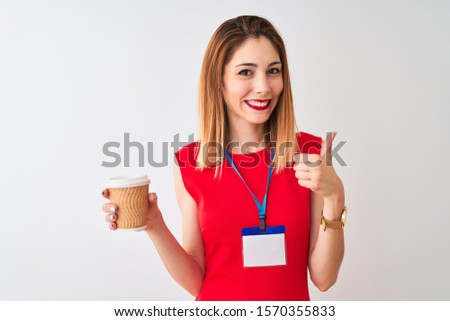 Redhead businesswoman wearing id card drinking cup of coffee over isolated white background happy with big smile doing ok sign, thumb up with fingers, excellent sign