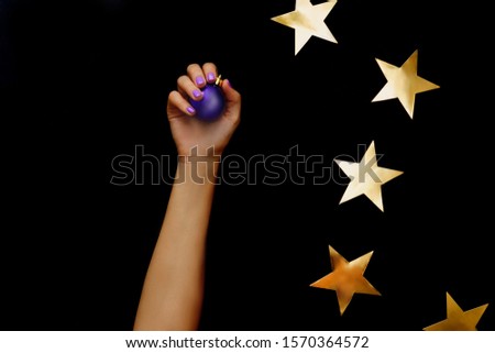 Beautiful christmas composition with gold stars. Violet christmas ball in hand with violet manicure on black background. Flat lay design.