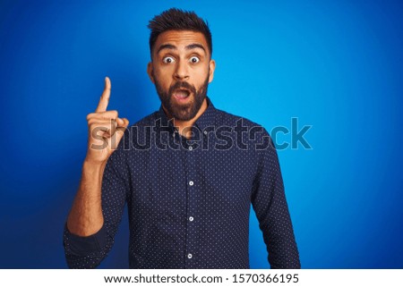 Young indian elegant man wearing shirt standing over isolated blue background pointing finger up with successful idea. Exited and happy. Number one.