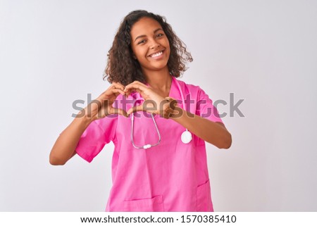 Young brazilian nurse woman wearing stethoscope standing over isolated white background smiling in love doing heart symbol shape with hands. Romantic concept.