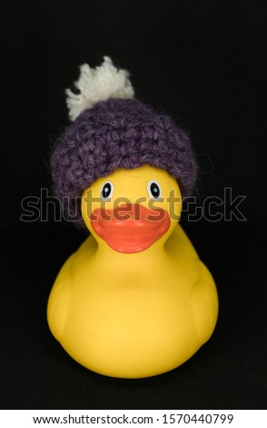 Funnly little toy. Yellow rubber duck with warm handmade purple wool hat with white pompom on a black background