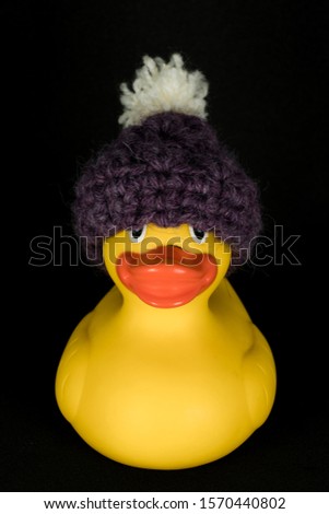 Funnly little toy. Yellow rubber duck with warm handmade purple wool hat with white pompom on a black background