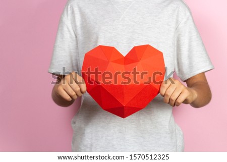Kid holding red polygonal paper 3d heart in his hands near light pink background