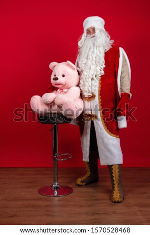 Emotional Santa Claus with a long white beard in a red coat and white hat posing with a big teddy pink bear on a red background