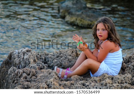 little baby girl with long, wavy hair looks at the sea sitting on a rock with a white dress and pink sandals
