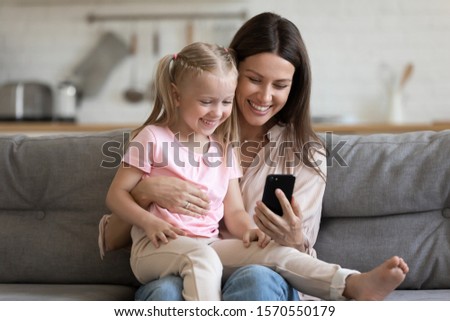 Happy family cute small kid child daughter sit on parent mom lap on sofa laugh having fun hold using smart phone watch funny social media video app take selfie bonding look at mobile screen at home