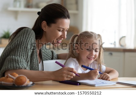 Young female babysitter mom teaching happy cute kid girl drawing with felt-tip pen together, smiling adult mother helping child daughter coloring picture enjoy creative activity at home sit at table