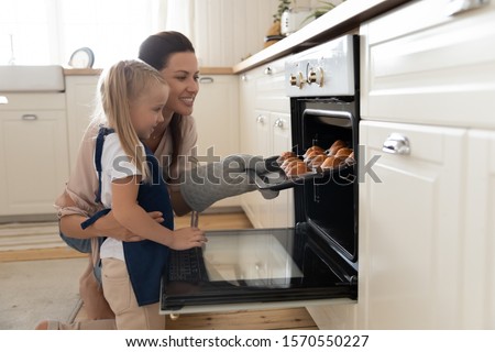 Happy young mum and cute kid daughter wear aprons bake muffins in kitchen, smiling mother teaching child take tray with cupcakes dessert make cakes out oven stove cook at home, family bakery concept