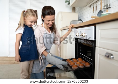 Excited cute small child daughter help mom bake muffins in kitchen oven, happy family mother with kid hold tray make cakes taking out ready homemade desserts out from stove having fun cook together