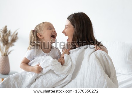 Happy cheerful family young adult mother having fun playing with adorable cute little kid child daughter holding warm white duvet laughing relaxing in cozy bed in morning enjoying family lifestyle