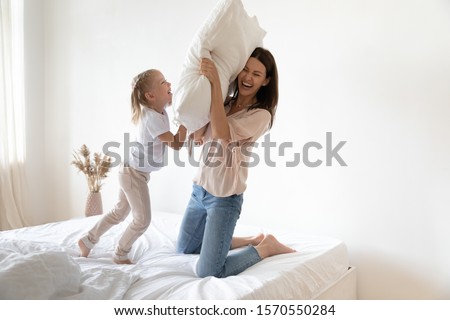 Happy active family young mother and cute small kid daughter laugh play pillow fight on cozy bed, carefree parent mum having fun with funny child at home bedroom enjoy morning leisure lifestyle game