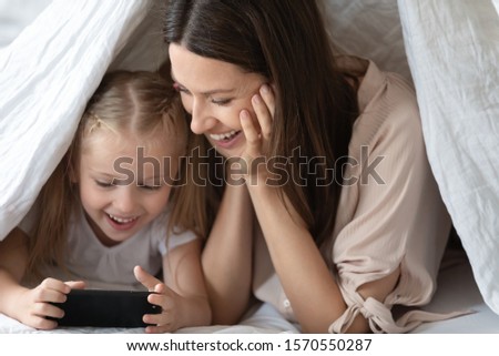 Happy family excited young adult mum and cute little kid child daughter lying on bed covered with blanket laugh having fun hold using smart phone play game app bonding look at mobile screen at home