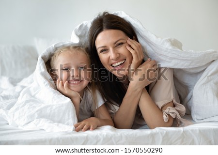 Happy family young mother and cute small kid daughter laugh look at camera covered with warm white duvet, smiling mom having fun with funny little child girl lay in cozy bed under blanket, portrait