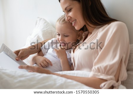 Caring single mama hug cute sweet kid daughter read book bedtime story lying in cozy bed in morning, happy loving family mother and child girl enjoying educational hobby at home bedroom together