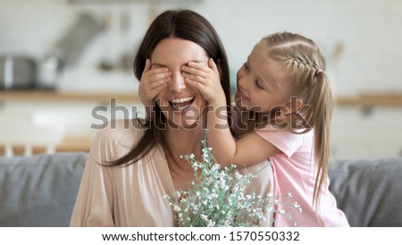 Adorable preschool child daughter make surprise close eyes of excited mommy presenting flowers at home, happy young mom receive bouquet from cute small kid girl celebrate mothers day together concept