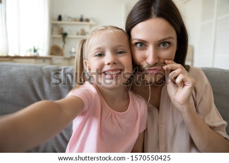Cute little kid daughter and young mother funny moustache face taking family picture look at mobile camera, happy parent mum having fun with child girl make selfie together at home, phone cam view Royalty-Free Stock Photo #1570550425