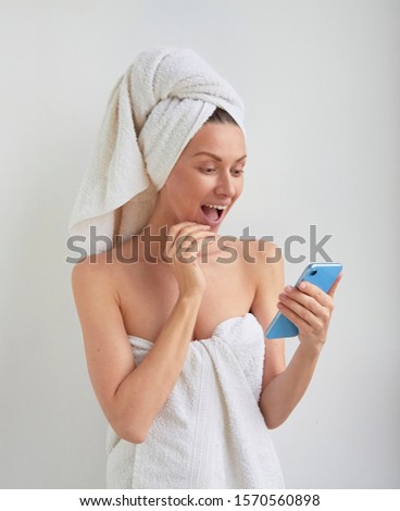Young beautiful woman with happy wondered surprised excited smile face wrapped hair on head and body in shower bath spa towel looks reading to the phone in hand on white room background Royalty-Free Stock Photo #1570560898