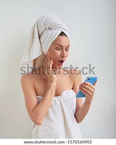 Young beautiful woman with happy wondered surprised excited smile face wrapped hair on head and body in shower bath spa towel looks reading to the phone in hand on white room background Royalty-Free Stock Photo #1570560901