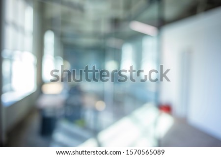 A blurry photograph of an office setting. The open corridor is flooded with natural light from the glass wall on the left. Blurred office interior space background Royalty-Free Stock Photo #1570565089